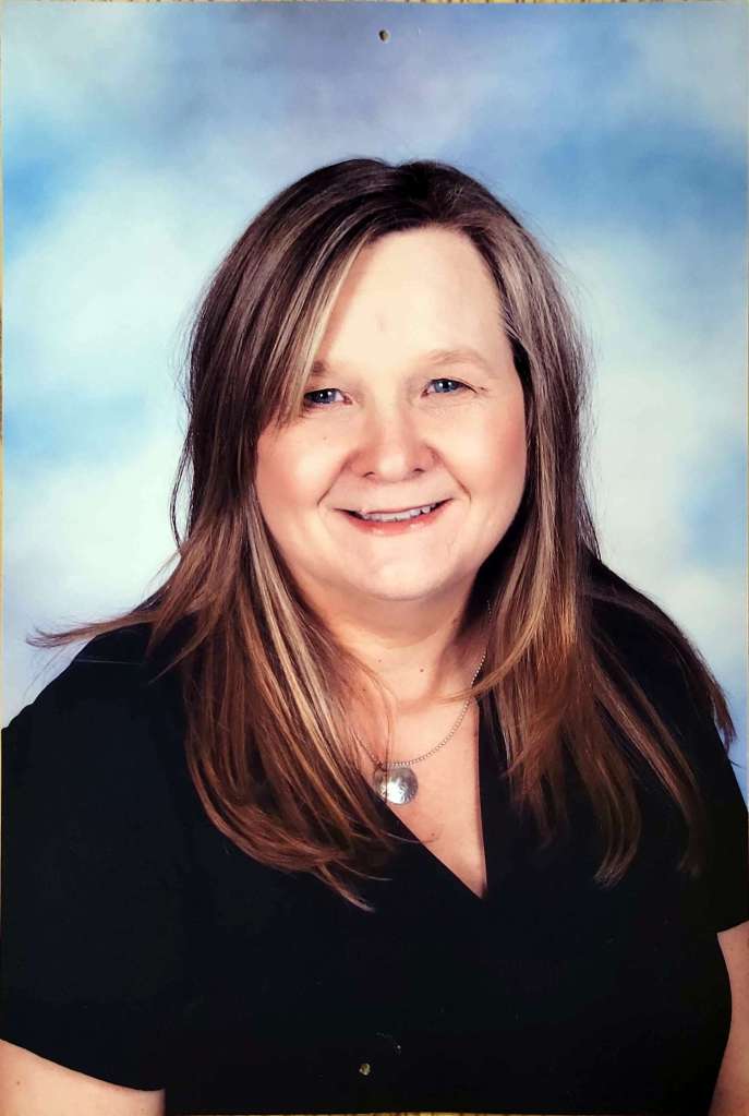This is a picture of Ms. Lori. Ms. Lori is a smiling woman with shoulder-length brown hair wearing a black top, and a silver necklace, set against a blue and white background.