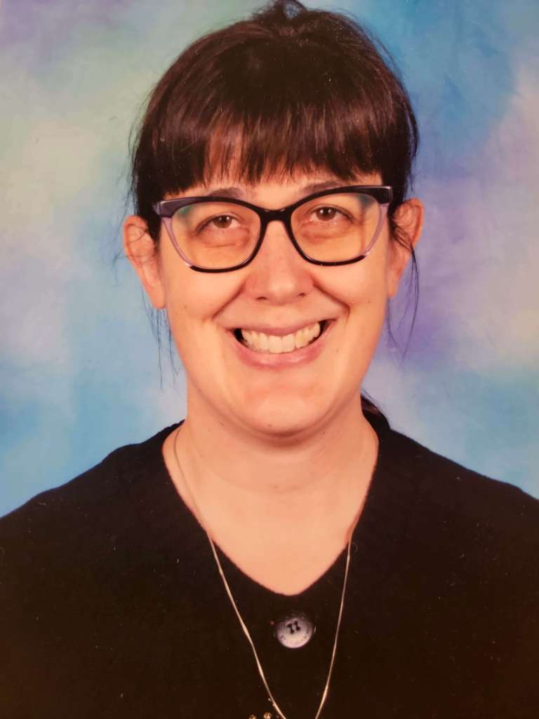 This is a picture of Ms. Kristin. Ms. Kristin is a smiling woman with dark hair pulled back in a ponytail with bangs. She is wearing dark framed glasses, a  black sweater, and is set against a light blue background.